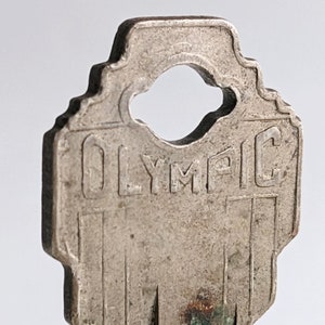 May include: Close-up of a silver key with the word "OLYMPIC" engraved on the top. The key has a unique shape with a large hole in the center.