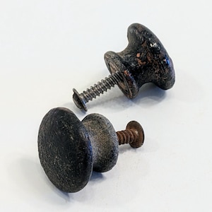 May include: Two black metal cabinet knobs with screws. The knobs have a slightly rounded, circular shape. One knob has a more worn, aged look.