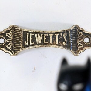 May include: A vintage metal sign with the text "JEWETT'S" in bold letters. The sign is rectangular with decorative ends and two holes for mounting. The sign has a worn, antique appearance.