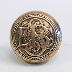 May include: A round, antique-style brass knob with a detailed, raised monogram of the letters "E", "B", and "S". The knob has a textured surface and a beaded border, suggesting a vintage or historical aesthetic.