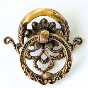 May include: A brass-toned metal drawer pull featuring an ornate, circular ring handle attached to a decorative backplate. The backplate displays intricate floral and leaf-like engravings, while the ring itself has a textured, patterned border.