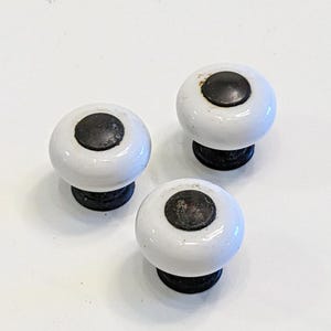 May include: Three white ceramic cabinet knobs with black centers. The knobs have a rounded, slightly domed shape.