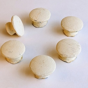 May include: Six off-white wooden knobs with a distressed finish. The knobs are round and have a slight raised edge. They are arranged in a circle on a white background.