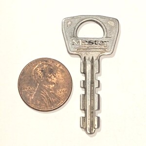 May include: A silver-colored metal key with the word Master embossed on the head, positioned next to a copper-colored United States one-cent coin for scale. The key features a flat, trapezoidal head with a circular cutout and a notched blade.