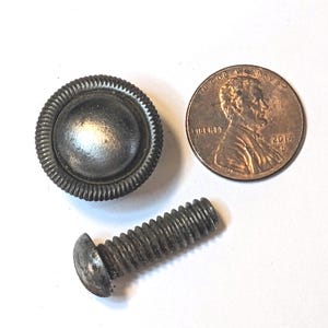 May include: A metallic, circular furniture knob with a ribbed edge and a smooth, domed center, accompanied by a threaded metal bolt and a copper-colored United States one-cent coin for scale, showing the profile of Abraham Lincoln and the year 2014.