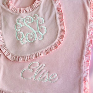May include: A pink baby bib and blanket set with ruffled edges. The bib features a monogram in white thread, while the blanket is embroidered with the name "Elise". The set is made of soft material, perfect for a baby.