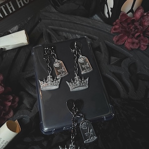 May include: A set of three silver-toned metal charms featuring a rose encased in a glass dome shape attached to a crown base. Two charms are connected to a black metal link chain, while the third is attached to a black heart-shaped metal piece.