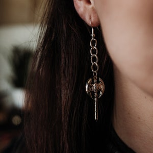 May include: A silver-colored earring featuring a small axe pendant suspended from a chain. The axe has intricate details and is attached to a chain of interconnected links. The earring is worn on the ear, with the hook visible.