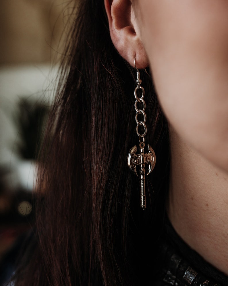 May include: A silver-colored earring featuring a small axe pendant suspended from a chain. The axe has intricate details and is attached to a chain of interconnected links. The earring is worn on the ear, with the hook visible.