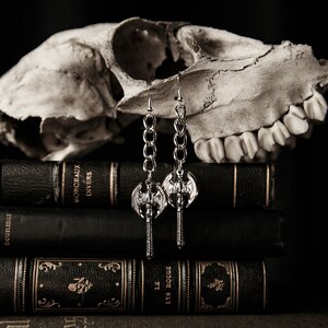May include: Silver-toned axe earrings with chain links hang in front of a skull and a stack of antique books. The earrings feature a double-bladed axe design. The books are dark with gold lettering and decorative elements.