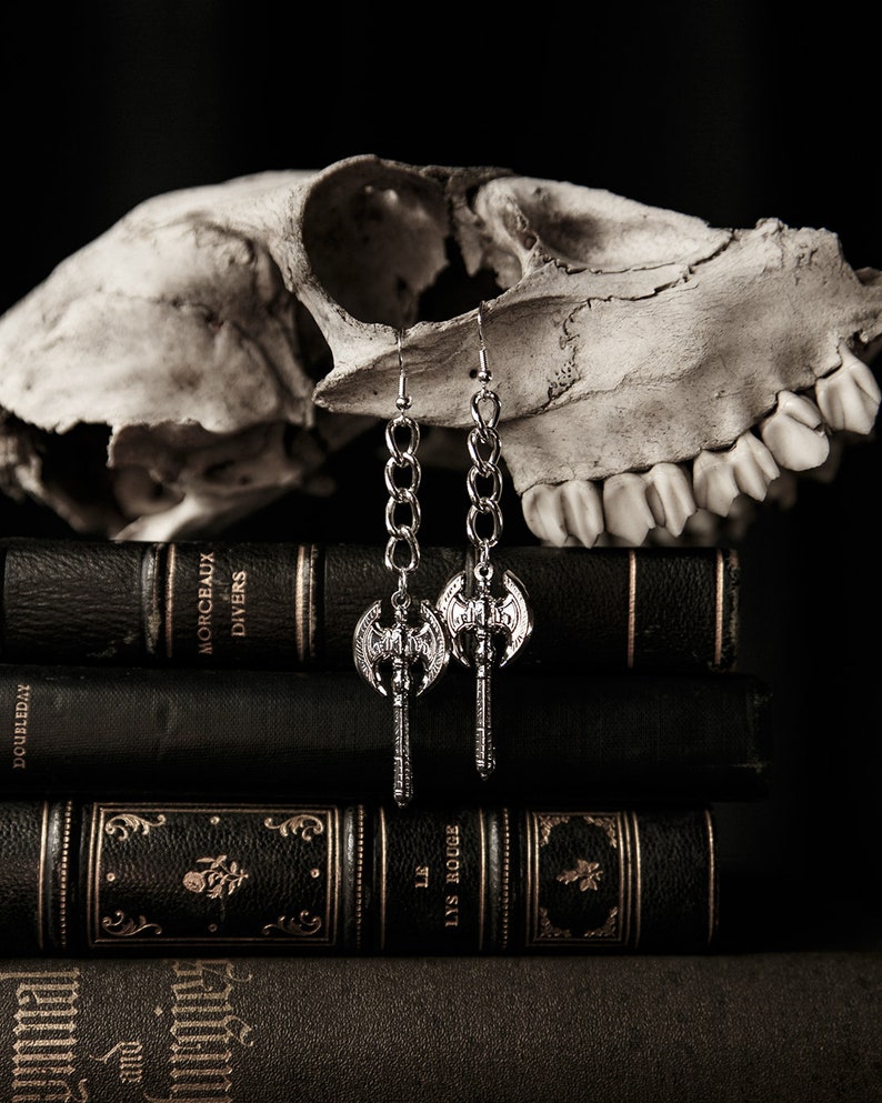 May include: Silver-toned axe earrings with chain links hang in front of a skull and a stack of antique books. The earrings feature a double-bladed axe design. The books are dark with gold lettering and decorative elements.