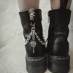Puede incluir: Botas de cuero negro con una cadena plateada y un dije de llave. Las botas tienen una suela de plataforma gruesa y una cremallera en el lateral. Las botas tienen una etiqueta amarilla que dice "AirWair" en la parte posterior.