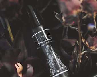 May include: A black essential oil roller bottle with the text "The Pretty Co. THE EMPRESS Blood Orange + Rosemary" and a white illustration of a woman. The bottle is surrounded by dark red and purple dried flowers.