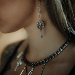 May include: Silver-toned axe earring hanging from a chain. A silver chain necklace with multiple charms, including a sword, is also visible. The jewelry has a gothic or fantasy theme.