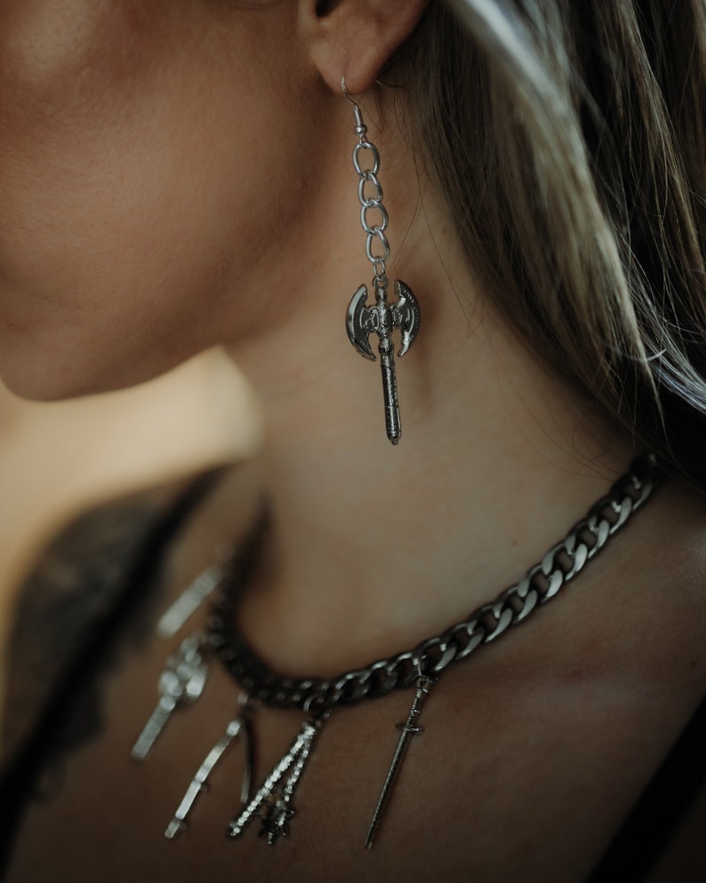 May include: Silver-toned axe earring hanging from a chain. A silver chain necklace with multiple charms, including a sword, is also visible. The jewelry has a gothic or fantasy theme.