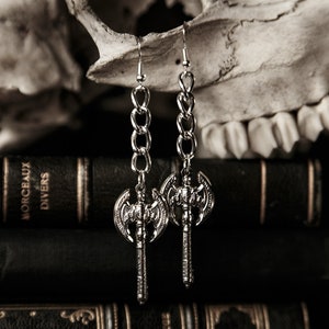 May include: Silver-toned axe earrings with chain links, displayed against a backdrop of antique books and a skull. The earrings feature detailed axe heads and long, dangling chains, creating a gothic aesthetic.