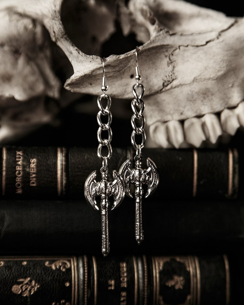 May include: Silver-toned axe earrings with chain links, displayed against a backdrop of antique books and a skull. The earrings feature detailed axe heads and long, dangling chains, creating a gothic aesthetic.
