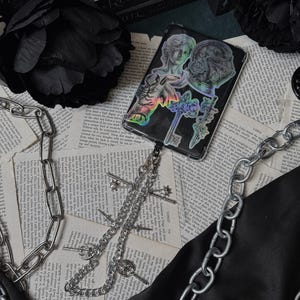 May include: A black phone case with holographic stickers of classical figures, keys, and a sword. A silver chain with key charms and a larger chain are draped over a black satin cloth and vintage book pages. Black roses and books are in the background.