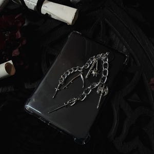 May include: A clear phone case with a silver chain and charms. The charms include a scythe, a cross, a dagger, and an axe. The background is dark, with rolled-up paper and the text "Ninth House".