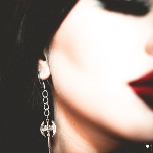 May include: Silver-toned axe-shaped earring with a chain detail. The earring features a double-bladed axe pendant with intricate detailing. The earring is worn on the ear, with a blurred background.