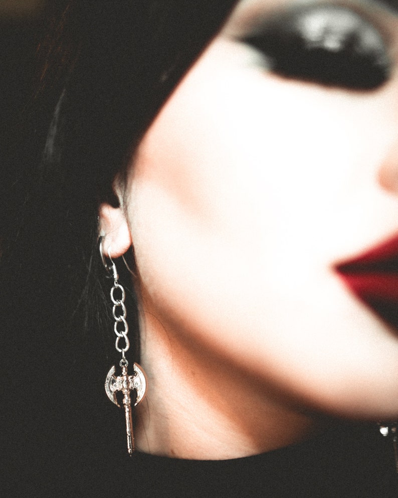 May include: Silver-toned axe-shaped earring with a chain detail. The earring features a double-bladed axe pendant with intricate detailing. The earring is worn on the ear, with a blurred background.