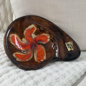 May include: A brown ceramic ashtray with a vibrant orange and cream floral design. The ashtray has a unique, organic shape and a glossy finish. A small label is visible on the side. The ashtray is a decorative item.
