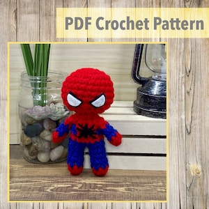 Puede incluir: Un juguete de amigurumi tejido a crochet de Spider-Man. El juguete es rojo y azul con un diseño de araña negra en el pecho. El juguete tiene una cara blanca con ojos negros y una boca roja.