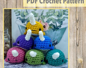 May include: A stack of five crocheted narwhal amigurumi toys in different colors, including yellow, purple, blue, pink, and green. The toys are arranged in a pyramid shape on a wooden surface.  PDF Crochet Pattern.