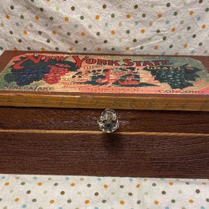 Könnte beinhalten: Eine Holzkiste mit einem Scharnierdeckel und einem Metallknopf. Der Deckel zeigt ein Vintage-Etikett mit dem Text "New York State Grapes" und Bildern von Trauben. Die Kiste ist braun mit einer Holzmaserung.