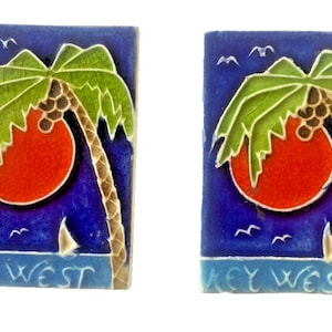 Könnte beinhalten: Zwei rechteckige Fliesen mit einem Key West-Design. Jede Fliese zeigt eine Palme, eine rote Sonne und ein Segelboot vor blauem Hintergrund. Die Worte "KEY WEST" stehen unten.
