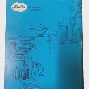 Puede incluir: Una portada de libro de cocina vintage con un fondo verde azulado e ilustraciones en blanco y negro de alimentos y artículos de cocina. El logotipo de Sunbeam está en la esquina superior izquierda, con el texto "built with integrity backed by service."