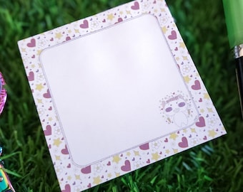 May include: A white card with a pink, yellow, and white border featuring hearts and stars. The card has a blank space in the center for writing a message. There is a small illustration of two cartoon animals in the bottom right corner.