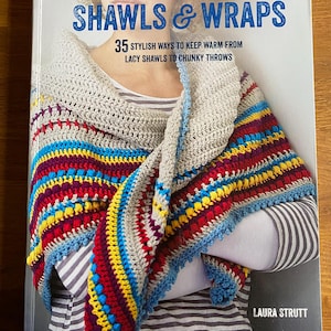May include: A book titled "Modern Crocheted Shawls & Wraps" featuring a colorful crocheted shawl. The shawl has a gray base with stripes of red, yellow, blue, and purple. The book cover also includes the text "35 Stylish Ways to Keep Warm From Lacy Shawls to Chunky Throws."