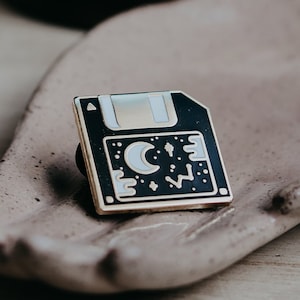Op de afbeelding: Een zwarte en gouden emaille pin in de vorm van een floppy disk met een halve maan en sterren op een zwarte achtergrond.