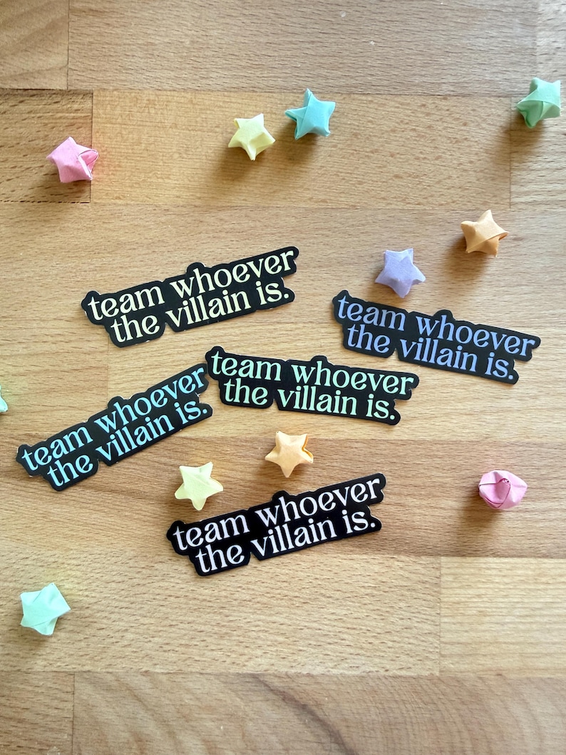 May include: Several black stickers with the text "team whoever the villain is" are scattered on a wooden surface, along with origami star decorations in various pastel colors. The stickers have a white border.