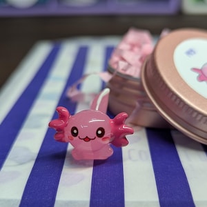 Op de afbeelding: Een roze axolotl-beeldje met een lachend gezicht en kleine oren, geplaatst op een blauw-wit gestreept oppervlak. Een roségouden blik met een roze axolotl-afbeelding staat op de achtergrond, samen met een kleine container gevuld met roze vloeipapier.