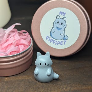 Op de afbeelding: Een klein grijs kattenbeeldje met een wit gezicht en een roze neus. Het beeldje staat op een houten oppervlak. Het beeldje staat voor een roze blik met een witte sticker waarop "My PippiPet" staat.