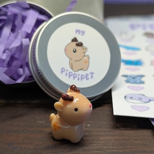 May include: A miniature deer figurine in tan with brown antlers and a pink nose. Beside it is a small silver tin with a label showing a cartoon deer and the text "MY PIPPIPET". Purple paper shreds are also visible.