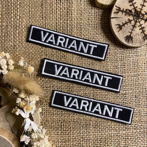 Peut inclure: Trois patchs en tissu noir avec du texte brodé blanc qui dit "VARIANT". Les patchs sont sur un fond de toile de jute brune.