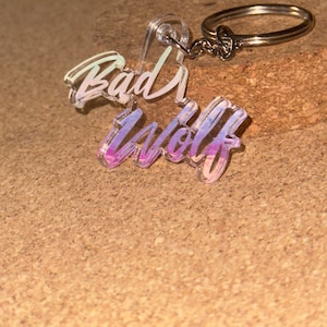 Peut inclure: Un porte-clés en acrylique transparent avec l'inscription "Bad Wolf" en écriture cursive. Le texte présente un effet dégradé, passant du violet au rose. Le porte-clés comprend un anneau et une chaîne argentés.