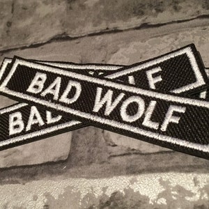 K&ouml;nnte beinhalten: Zwei schwarze Stoff-Patches mit wei&szlig; gesticktem Text, der "BAD WOLF" lautet.