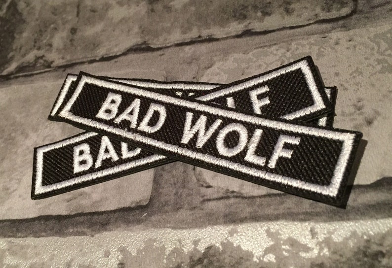 K&ouml;nnte beinhalten: Zwei schwarze Stoff-Patches mit wei&szlig; gesticktem Text, der "BAD WOLF" lautet.