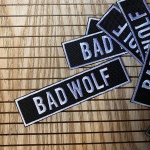 May include: Black and white embroidered patches with the words "BAD" and "BAD WOLF" on a wooden background.
