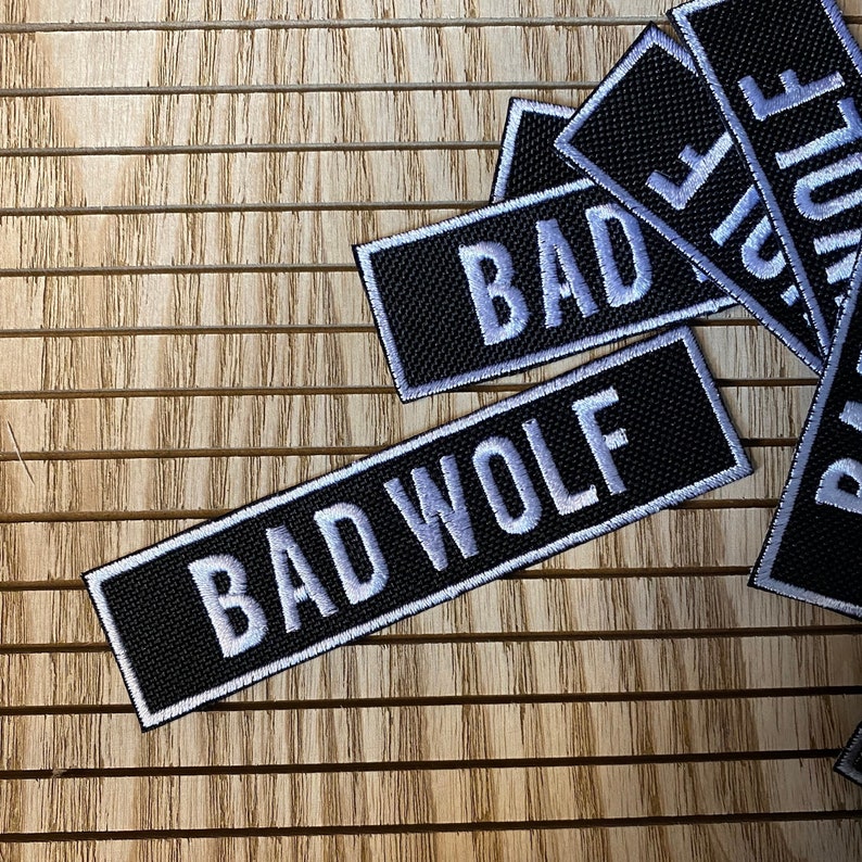 K&ouml;nnte beinhalten: Schwarze und wei&szlig;e gestickte Patches mit den Worten "BAD" und "BAD WOLF" auf einem Holzgrund.