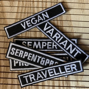 May include: A stack of black and white embroidered patches with the words "Vegan", "Variant", "Semper", "Serpent & Pride", "Traveler" in white lettering.