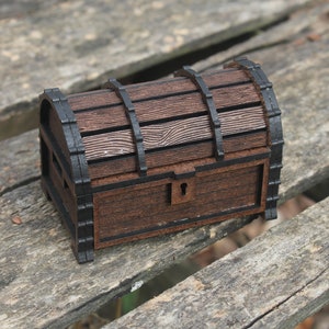 Puede incluir: Un pequeño cofre del tesoro de madera con una tapa con bisagras y una cerradura de metal. El cofre está pintado de marrón con detalles negros y tiene un patrón de veta de madera.