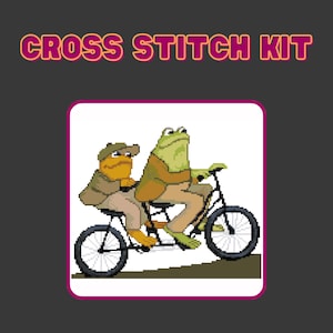 Könnte beinhalten: Ein Kreuzstich-Set mit einer pixeligen Illustration von zwei Fröschen auf einem Tandem-Fahrrad. Die Frösche tragen Hüte und Jacken. Der Text "CROSS STITCH KIT" steht in Rosa und Gelb über dem Bild.