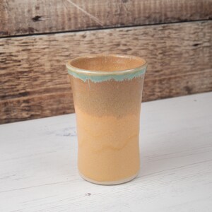 May include: A ceramic mug with a light brown glaze and a light blue rim. The mug is sitting on a white surface.