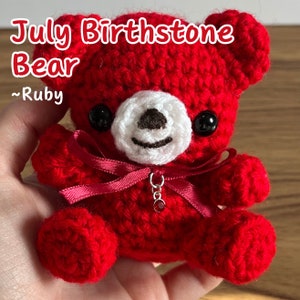 May include: A red crocheted teddy bear with a white face and black eyes. The bear is wearing a red ribbon and a charm with a red gemstone. The text "July Birthstone Bear ~ Ruby" is written above the bear.