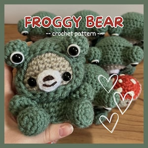 Puede incluir: Un disfraz de rana de crochet para un osito de peluche. El oso lleva un disfraz de rana verde con grandes ojos saltones. El oso está sentado sobre una superficie de madera con otros animales de crochet.